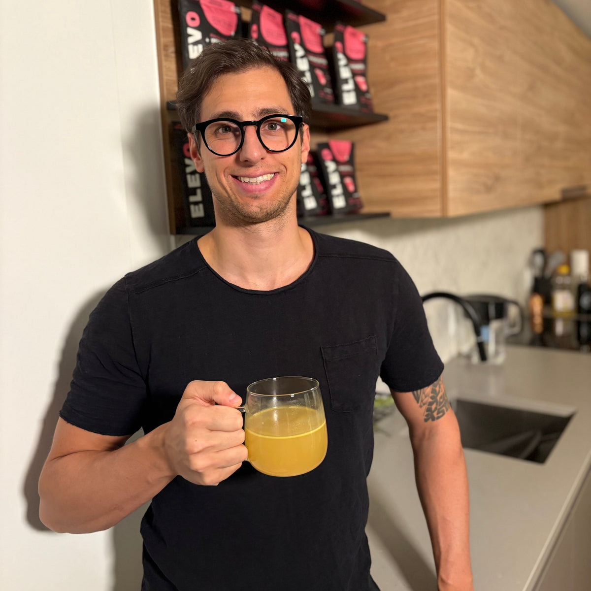Man holding a mug of juice in a kitchen with shelves displaying ELEVO Nutrition products.
