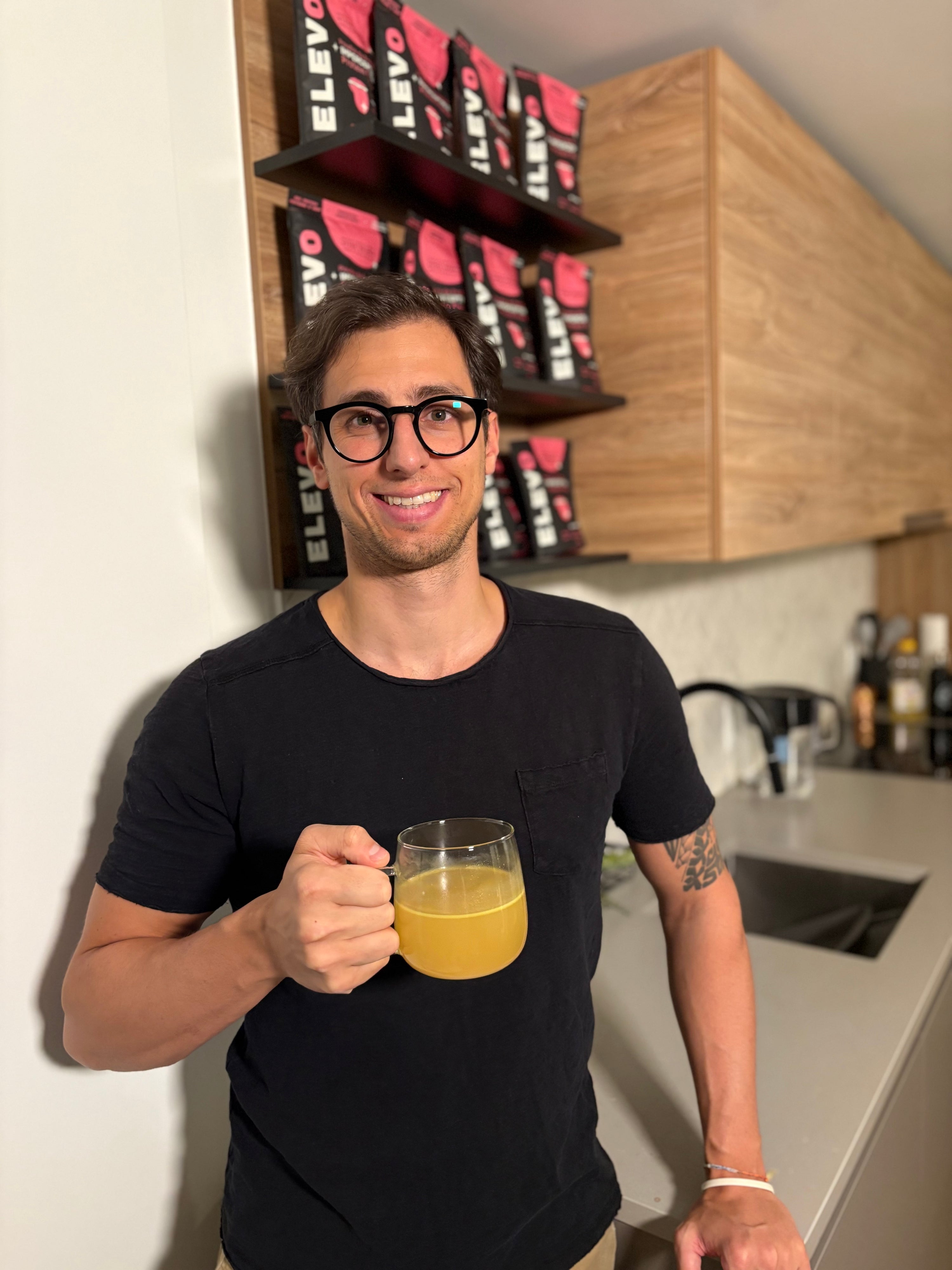 Man holding a mug of juice in a kitchen with shelves displaying ELEVO Nutrition products.