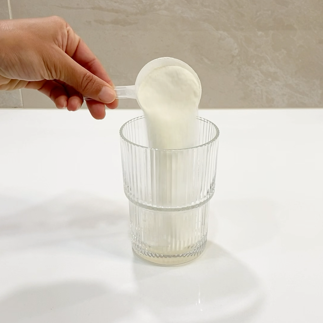 Hand holding a spoon with a scoop of ELEVO Nutrition protein powder over a clear glass