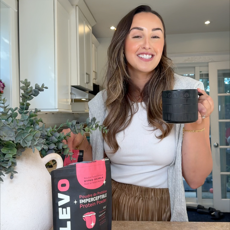 Woman holding a black mug and a bag of ELEVO protein powder in a kitchen.